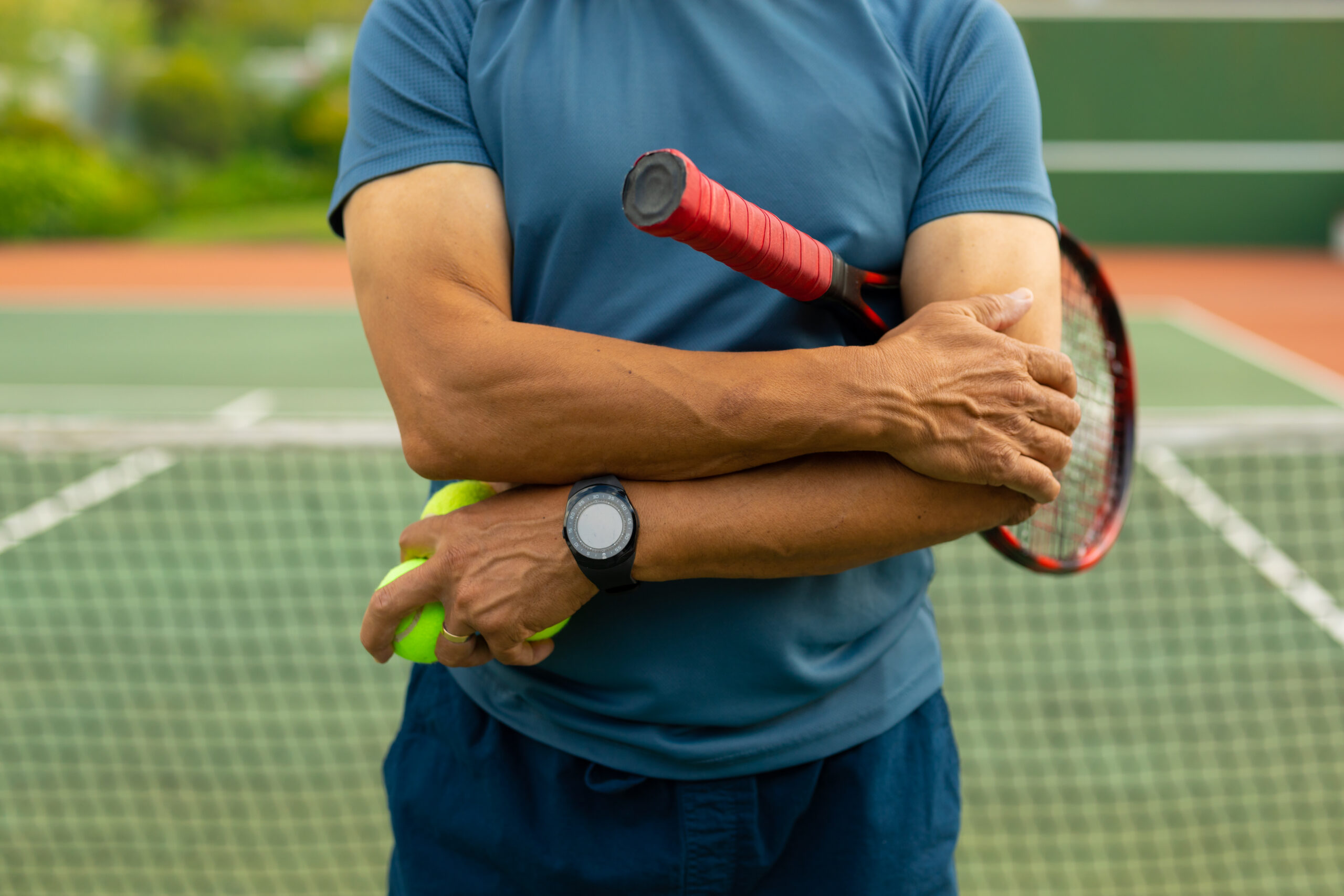 Oberkörper eines mittelalten Mannes auf einem Tennisplatz, der sich mit der rechten Hand den linken schmerzenden Ellenbogen hält - Akupunktur bei Tennisarm - Grollmann-TCM